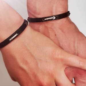 Op de afbeelding: Twee zwarte leren armbanden met zilveren pijlpuntaccenten. De armbanden worden gedragen op de polsen van twee mensen die elkaars handen vasthouden.