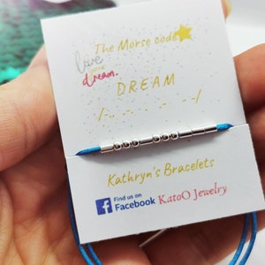 Può includere: Un braccialetto in corda blu con perline d'argento che compongono la parola "DREAM" in codice Morse. La carta recita "Il codice Morse per sognare. DREAM" e "Braccialetti di Kathryn Trovaci su Facebook KatoO Jewelry".