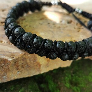 Puede incluir: Pulsera de piedra de lava negra con cord&oacute;n negro ajustable. La pulsera presenta cuentas de lava porosas y redondas ensartadas con un cord&oacute;n negro tejido. El cierre ajustable tiene dos peque&ntilde;as cuentas de color plateado. La pulsera se muestra sobre una superficie de piedra marr&oacute;n claro.