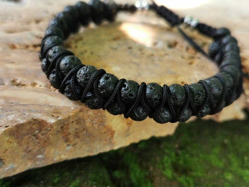 Puede incluir: Pulsera de piedra de lava negra con cord&oacute;n negro ajustable. La pulsera presenta cuentas de lava porosas y redondas ensartadas con un cord&oacute;n negro tejido. El cierre ajustable tiene dos peque&ntilde;as cuentas de color plateado. La pulsera se muestra sobre una superficie de piedra marr&oacute;n claro.