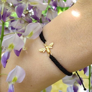May include: A black cord bracelet with a gold bee charm. The bracelet is worn on a wrist against a background of purple and white wisteria flowers.