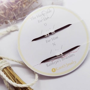 May include: Two black string bracelets with silver beads, one with the Morse code for "U" and the other with the Morse code for "N". The bracelets are on a white card with the text "The Morse code For Her U For Him N Kathryn's Bracelets Katoo Jewelry".