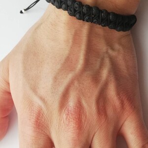 Puede incluir: Pulsera de roca volc&aacute;nica negra con cord&oacute;n negro ajustable. La pulsera presenta una doble hebra de cuentas porosas de color gris oscuro. El cord&oacute;n est&aacute; atado con una peque&ntilde;a cuenta blanca en el extremo. La pulsera se lleva en la mu&ntilde;eca.