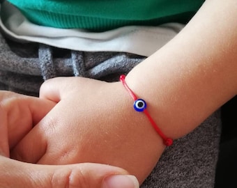 May include: A red string bracelet with a blue evil eye charm. The bracelet is worn on a child's wrist.