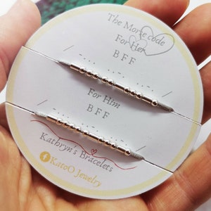 May include: A white card with the text "The Morse code" and two heart symbols. The card also has the text "For Her BFF" and "For Him BFF" with two silver bracelets with beads and a silver bar. The card also has the text "Kathryn's Bracelets Katoo Jewelry".