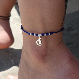 May include: A blue anklet with a silver sailboat charm. The anklet features small silver beads and is worn on an ankle. The charm is a detailed sailboat design, adding a nautical touch to the accessory.