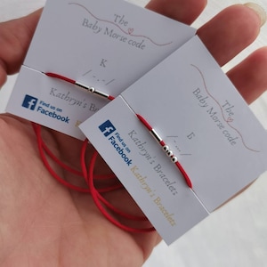 May include: Two red string bracelets with silver beads and a silver clasp. The bracelets are on cards with the text "The Baby Morse code" and the letter "K" and "B" in Morse code. The cards also have the text "Kathryn's Bracelets" and a Facebook logo.