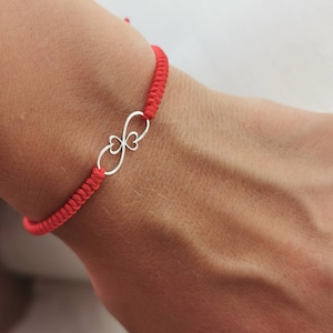 Può includere: Un braccialetto di corda rossa con un ciondolo d'argento a forma di simbolo di infinito con due cuori.