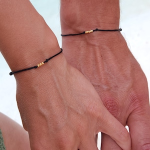 Può includere: Due braccialetti neri in corda con perline dorate. I braccialetti sono indossati sui polsi di due persone che si tengono per mano.