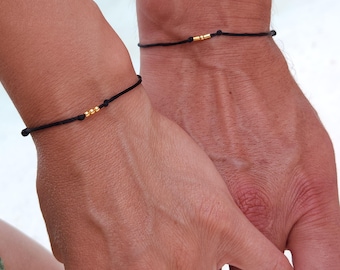 Puede incluir: Dos pulseras de cuerda negra con cuentas doradas. Las pulseras se llevan en las muñecas de dos personas que se toman de la mano.