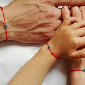 May include: Three red string bracelets with blue beads, worn on the wrists of three people, showing the hands of an adult and two children.