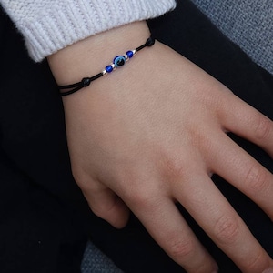Puede incluir: Un brazalete de cuerda negra con un dije azul de ojo turco. El dije tiene un borde plateado y detalles blancos.