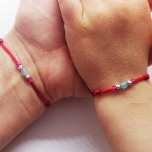 Puede incluir: Dos pulseras de cuerda roja con cuentas plateadas y cuentas de piedra preciosa azul claro. Las pulseras están en las muñecas de dos personas.