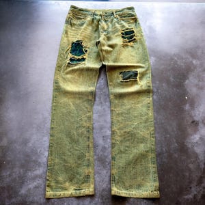Può includere: Jeans in denim verde oliva slavato con più sezioni strappate che rivelano un tessuto verde più scuro sottostante. I jeans hanno un aspetto sbiadito e usurato e un classico design a cinque tasche. I jeans sono appoggiati su una superficie scura.