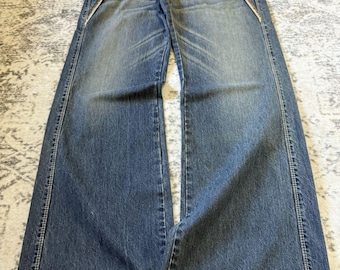 May include: Blue denim jeans with a wide leg design. The jeans feature a faded wash, visible stitching, and decorative zippers along the front pockets. The waistband is a lighter tan color. The jeans are laid flat on a patterned surface.