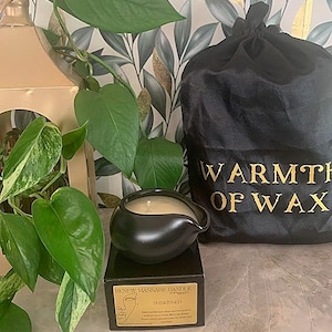 May include: A black massage candle in a unique, rounded shape with a spout, sitting on a black box. The box has text that reads "Renew Massage Candle" and "Sunkissed". A black bag with gold text "Warmth of Wax" is in the background.