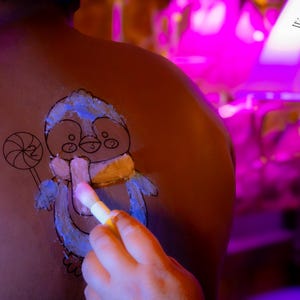 May include: A person is getting a blue and white penguin painted on their back with a white paintbrush. The penguin is holding a lollipop. The background is purple and pink.