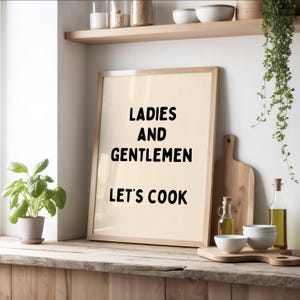 Peut inclure: Un cadre en bois clair avec l'inscription en noir « LADIES AND GENTLEMEN LET'S COOK ». Il est posé contre une planche à découper en bois. On aperçoit également des bols, des bouteilles d'huile et une plante en pot.