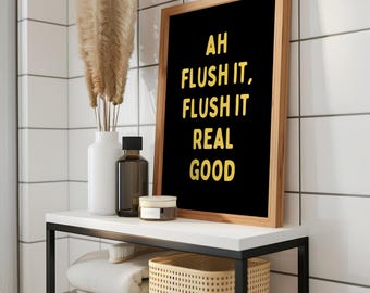 May include: A bathroom shelf with a black metal frame and white top holds a framed black sign with yellow text that reads "AH FLUSH IT, FLUSH IT REAL GOOD." Other items include a white vase with dried plants, a brown bottle, and a small jar.