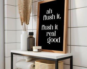 May include: A bathroom shelf with a black metal frame and white top holds a framed black print with white text that reads "ah flush it, flush it real good." A white vase with dried flowers, a brown bottle, and a small jar are also on the shelf.