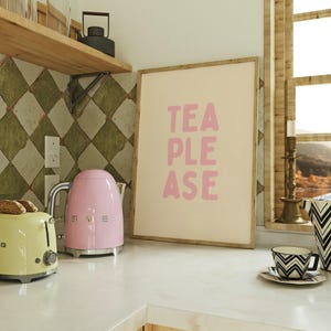 May include: A pink electric kettle and a yellow toaster on a white countertop with a framed print that says "TEA PLEASE" in pink letters on a beige background.