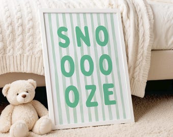 May include: A framed print with the word "SNOOOZE" in green letters on a striped background. A white teddy bear sits on a cream-colored rug next to the framed print. The background includes a bed with a white blanket.