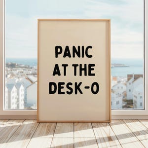 May include: A framed print with a beige background and black text that reads "PANIC AT THE DESK-O".