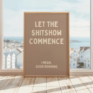 Könnte beinhalten: Ein gerahmter Druck mit dem Text "LET THE SHITSHOW COMMENCE" in einer fetten, cremefarbenen Schrift. Darunter steht in kleinerer Schrift "I MEAN, GOOD MORNING". Der Rahmen ist hellbraun, der Hintergrund ist ein gedämpftes Braun.