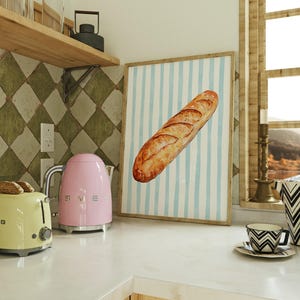 May include: A kitchen setting featuring a framed print of a baguette against a blue and white striped backdrop. A pink electric kettle and a yellow toaster are on the counter. A black and white patterned cup and vase are also visible.