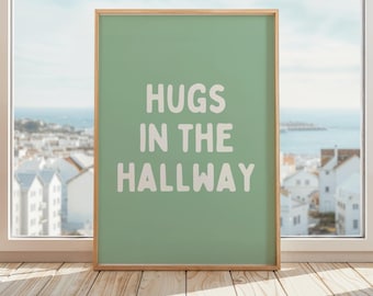 May include: A framed art print with the words "HUGS IN THE HALLWAY" in white, bold letters on a light green background. The frame is a light wood color. The print is displayed on a wooden floor in front of a window with a view of a coastal town.