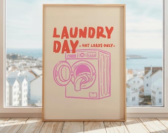 Op de afbeelding: Een ingelijste print met de tekst "LAUNDRY DAY - HOT LOADS ONLY" in het rood en een roze illustratie van een wasmachine met een open deur. De achtergrond is lichtbeige en de lijst is lichtbruin.