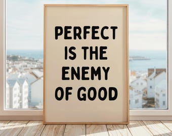 May include: A framed print with the text "PERFECT IS THE ENEMY OF GOOD" in bold black letters. The print has a cream-colored background and a light wood frame. The print is displayed on a wooden floor in front of a window.