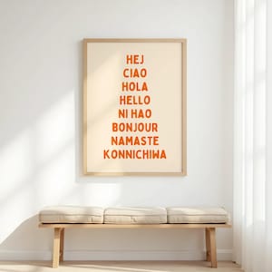 Może przedstawiać: Oprawiony wydruk artystyczny ze słowami "Hej, Ciao, Hola, Hello, Ni Hao, Bonjour, Namaste, Konnichiwa" w pomarańczowych literach. Wydruk jest w jasnej drewnianej ramie i wisi nad ławką z trzema beżowymi poduszkami.