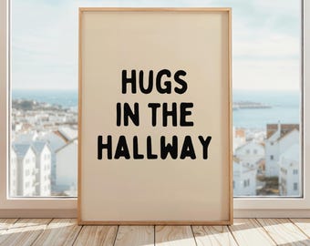 May include: A framed print with the words "HUGS IN THE HALLWAY" in bold black letters. The print has a cream-colored background and a light wood frame. It is placed on a wooden floor in front of a window overlooking a coastal town.