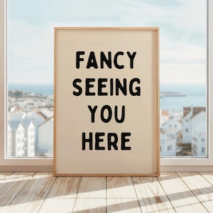 Può includere: Una stampa artistica incorniciata con la scritta "FANCY SEEING YOU HERE" in grassetto nero. La stampa ha uno sfondo color crema e una cornice in legno chiaro. La stampa è esposta davanti a una finestra che si affaccia su una città costiera.