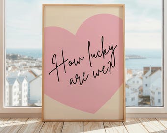 May include: A framed art print featuring a light pink heart shape on a cream background. The black cursive text reads "How lucky are we?" The artwork is displayed on a wooden surface in front of a window with a coastal view.