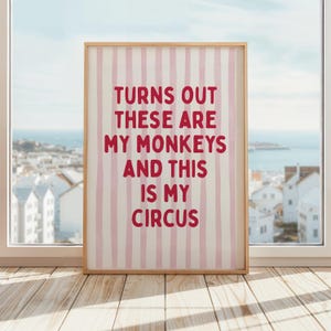 Può includere: Una stampa incorniciata con una cornice in legno chiaro. Lo sfondo presenta strisce verticali rosa e bianche. Il testo rosso in grassetto recita: "TURNS OUT THESE ARE MY MONKEYS AND THIS IS MY CIRCUS."