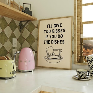 May include: A framed print with the text "I'LL GIVE YOU KISSES IF YOU DO THE DISHES" and a dish illustration. A pink electric kettle and a yellow toaster are on the counter. A black and white patterned cup and saucer are also visible.
