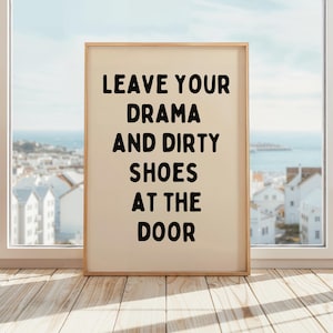 Peut inclure: Une affiche encadrée avec le texte "LEAVE YOUR DRAMA AND DIRTY SHOES AT THE DOOR" en lettres noires grasses. L'affiche a un fond crème et un cadre en bois clair. L'affiche est affichée sur un sol en bois devant une fenêtre.