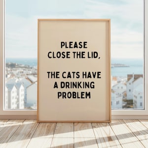 Può includere: Una stampa incorniciata con la scritta "PLEASE CLOSE THE LID, THE CATS HAVE A DRINKING PROBLEM" in lettere nere. La stampa è in una cornice di legno chiaro e si trova su un pavimento di legno davanti a una finestra con vista su una città costiera.