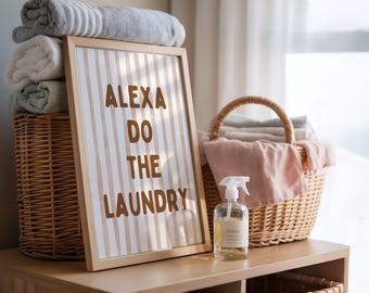 May include: A wooden framed sign with the text "ALEXA DO THE LAUNDRY" stands on a wooden shelf, next to a wicker basket filled with towels. A spray bottle and more towels are also visible. The sign has a striped background.