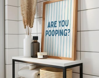 Op de afbeelding: Een badkamerplank met een ingelijste print met de tekst "ARE YOU POOPING?" in blauw op een gestreepte achtergrond. De plank bevat ook een witte vaas met gedroogde planten, een bruine fles, een klein potje, een gevlochten mand en opgevouwen handdoeken.