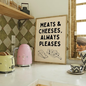 May include: A kitchen setting featuring a framed print with the text "MEATS & CHEESES, ALWAYS PLEASES." A pink electric kettle and a yellow toaster are on the counter. A patterned cup and saucer are also visible.