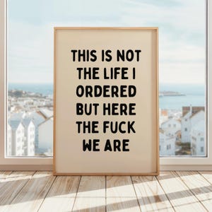 May include: A framed print with the text "THIS IS NOT THE LIFE I ORDERED BUT HERE THE FUCK WE ARE" in bold black letters. The print has a light beige background and a natural wood frame. It is displayed on a wooden surface in front of a window.