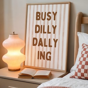 May include: A framed print with the words "BUSY DILLY DALLY ING" in brown letters on a striped background. A decorative lamp and an open book sit on a nightstand next to a bed with a patterned pillow.