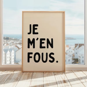 May include: A framed print featuring the French phrase "JE M'EN FOUS." in bold black lettering on a cream-colored background. The wooden frame is placed on a wooden floor, with a coastal view visible through a window.