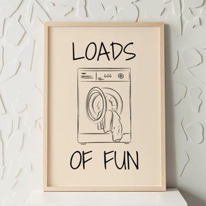 May include: A beige framed print with a black and white illustration of a washing machine with the text "LOADS OF FUN" in black.