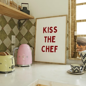 Op de afbeelding: Een keuken met een roze elektrische waterkoker en een gele broodrooster op een wit aanrecht. Een ingelijste print met de rode tekst "KISS THE CHEF" is te zien. Een kop en schotel met zwart-wit patroon zijn ook zichtbaar.
