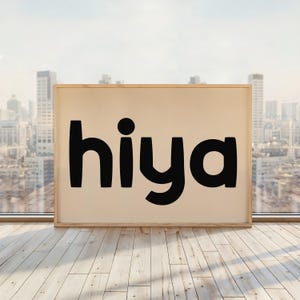 Könnte beinhalten: Gerahmter Druck mit dem Wort "hiya" in fetten, schwarzen Buchstaben. Der Hintergrund ist hellbeige, und der Rahmen ist aus hellem Holz. Der Druck wird auf einem Holzboden vor einem Fenster mit Blick auf die Skyline präsentiert.