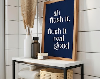 May include: A bathroom shelf displays a framed navy blue print with white text reading "ah flush it, flush it real good." A white vase with dried pampas grass, a brown bottle, and a small jar are also on the shelf. Below are folded towels and a woven basket.